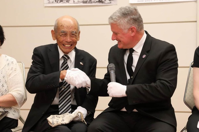 日章旗を手に笑顔で握手する陸田敏弘さん（左）とUSSレキシントン博物館のスティーブ・バンタ館長（右）