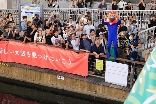 「トビコムナ」と書かれたプレートを掲げた「ミャクミャク」のコスプレをした人物。この後、川へ飛び込んだ（7日、大阪市中央区）