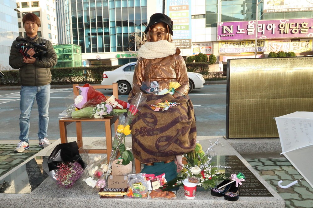釜山の日本総領事館前に設置された慰安婦像。ストールやマフラーが巻かれ、お菓子や花、靴が備えられている