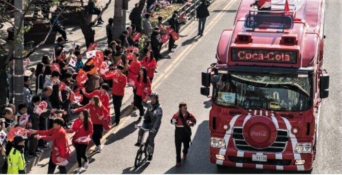 沿道でグッズを配る聖火リレーのスポンサー、コカ・コーラの車列（同社の公式サイトより）