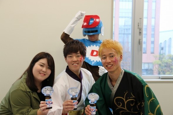 リポビタンDを贈る赤羽美香さん（左）と、関東学院大学「誇咲」の赤羽亮太さんと山﨑湧太さん。そして背後のヒーローは何者？